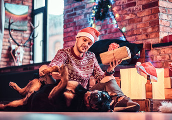 A stylish man holding a gift box while sitting with his cute dog in a decorated living room at Christmas time.