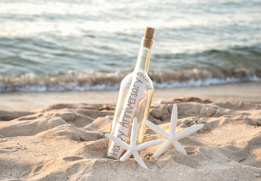 Pair Of White Starfish In Beach Sand With Happy Anniversary Message In A Bottle On The Seashore