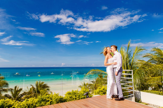 Beautiful blonde bride in white wedding dress and the groom on the roof of the hotel. Tropical sea and palm trees in the background. Summer vacation concept.