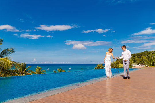Beautiful blonde bride in white wedding dress and the groom dance in a hotel near the infinity pool. Tropical sea, sky and palm trees in the background. Summer vacation concept.