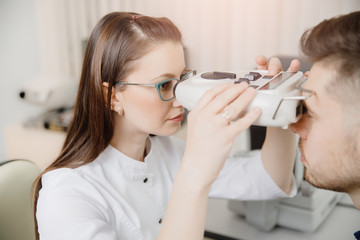 Attractive girl doctor ophthalmologist checks vision of young man with fashionable haircut and beard on special equipment