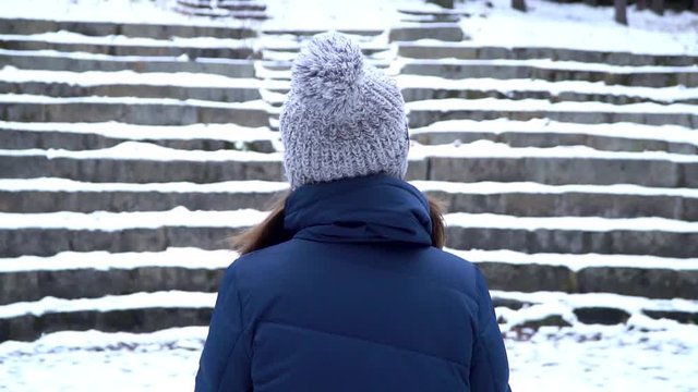 Rear view on woman with hat stays in a park while snowing, snowy stairs background. Alone woman stand alone. snow in winter, landscape, romantic scene.