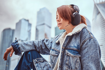 A hipster girl with tattoo on her face trendy dressed wearing headphones listening to music in front of skyscrapers in Moskow city at cloudy morning.