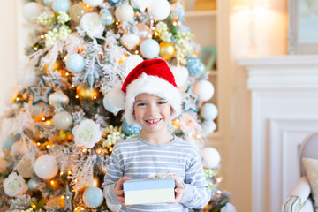 Happy smiling Boy in santa hat with gift box over christmas tree lights background. Christmas and New Year concept. Childghood.