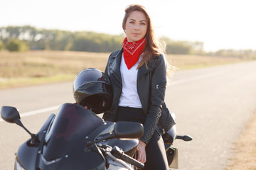 Outdoor horizontal shot of attractive woman wears red bandana, leather jacket, holds helmet, sits on motorrbike, has active lifestyle, being professional biker, enjoys speed. People and hobby concept
