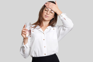 Tired overworked woman keeps hand on forehead, wears transparent glasses, black and white clothes,...