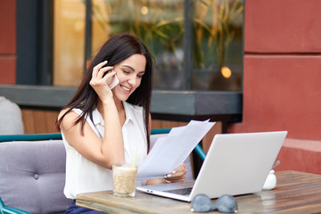 Obraz premium Wesoła brunetka bizneswoman prowadzi rozmowę telefoniczną, zadowolona z doradztwa, rozmawia z operatorem, ubrana w modną bluzkę, trzyma papierowe dokumenty, pracuje na laptopie