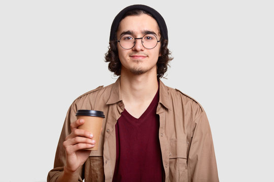 Handsome boy has curly hair, beard wears round transparent glasses, holds coffee to go, has break, communicates with friends, models against white background. People, lifestyle, drink concept