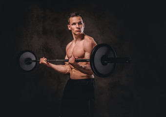 Young shirtless bodybuilder holding a barbell and doing exercise on biceps. Isolated on dark...
