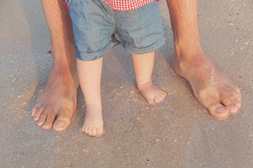 Man and baby feet standing in shallow water waiting for the wave. Bare feet father and his little daughter or son staying in the sand near the sea. Concept of travel and holidays. Toned. Soft focus.