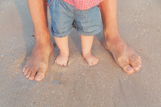 Man And Baby Feet Standing In Shallow Water Waiting For The Wave. Bare Feet Father And His Little Daughter Or Son Staying In The Sand Near The Sea. Concept Of Travel And Holidays. Toned. Soft Focus.