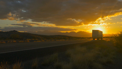 Freight semi truck speeding on empty highway over golden sun at summer sunset