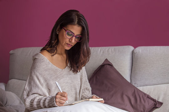 Mujer morena escribiendo a mano en el sof&aacute; en un cuaderno con bol&iacute;grafo y papel