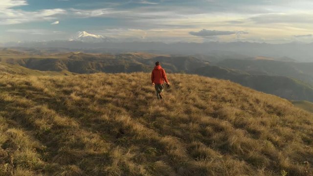 An aerial view of a bearded hipster male with a remote from a drone walks along the edge of a high plateau near a cliff at sunset. The guy walks along the cliff