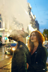 Couple in love on date. Boyfriend smokes electronic cigarette. Girlfriend hugs him. They walk streets of night city. Wet asphalt after rain.