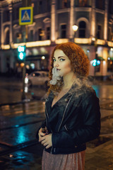 Young red-haired girl smokes an electronic cigarette. She is walking in street of night city. Wet...