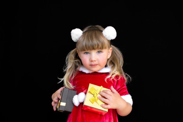 Little beautiful girl in a Christmas Santa suit, with fur balls on her head, holds gifts in her hands and rejoices. The concept of celebrating Christmas, New Year and gifts.