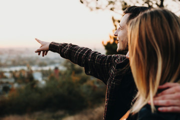 Side view of handsome young guy embracing girlfriend and pointing at distance while standing on blurred background of nature together. Couple looking at distance in nature