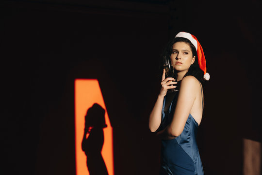 Side view of attractive young female in Christmas hat and elegant dress looking away and thinking while standing in dark room and holding glass of chamagne