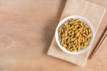 top view of roasted silkworm pupae with salt and soy sauce in a ceramic dish on wooden table, copy space. alternative high nutrition food concept.