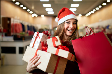 Happy girl shopping gifts in mall on christmas sale. New year holidays shopping idea concept. Smiling woman with colorful paper presents bags and gift boxes wearing christmas hat in store or shop.