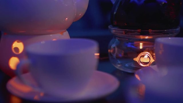 Focus shifting from teacup onto teapot with candle under it for heating on table in caffee