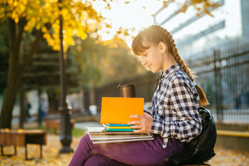 Happy 12 years old teen girl in eyeglasses with two braids reading workbooks in the autumn park after school.