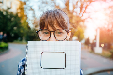 Cute 12 years old school girl in eyeglasses, plaid shirt holding text book covering half of her...
