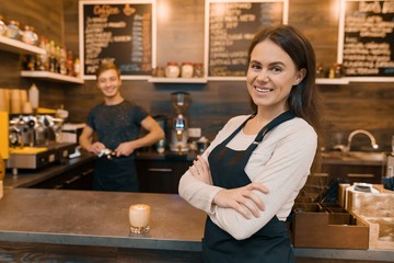 Portrait of smiling young female coffee shop owner, confident woman with arms crossed standing at the counter with barista working in background making drinks