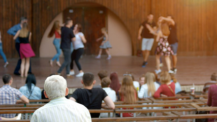 People at outdoor concert. Old man sitting on bench and looking at performance on the stage, back...