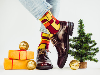 Men's legs, funny socks, stylish shoes, gifts with a red ribbon and a Christmas tree. White...