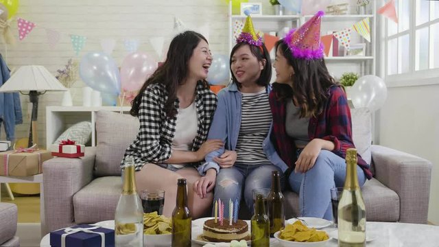 slow motion of happy party young girls sitting close on couch talking some funny things and laughing together. beautiful asian ladies with birthday hats sitting around the cake food drinks on table.