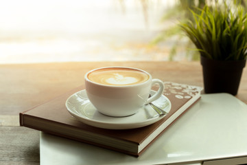 Cup of coffee on top of book and laptop on wooden table in the morning.
