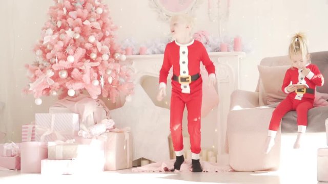 The boy blond is dancing at the New Year tree. His younger sister is sitting on the couch and playing on the phone. Albino dressed as Santa Claus jumps to the top. Children enjoy Christmas. Family