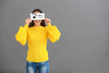 Emotional young woman hiding face behind sheet of paper with drawn eyes on grey background