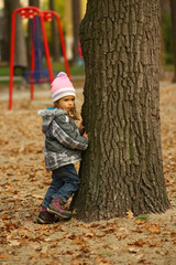 Toddler girl playing hide and seek in the autumn park and hiding behind the tree. Ful length portrait