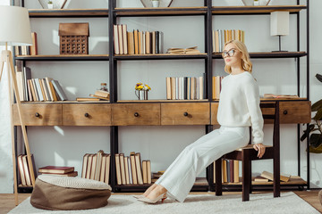 adult blonde woman in glasses sitting near rack with books