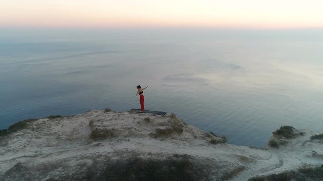 Beautiful freedom woman is doing yoga stretcing on the top of mountain with sea view at sunset. Aerial footage.