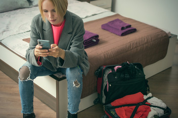 A woman is sitting with a phone on the bed near the suitcase.