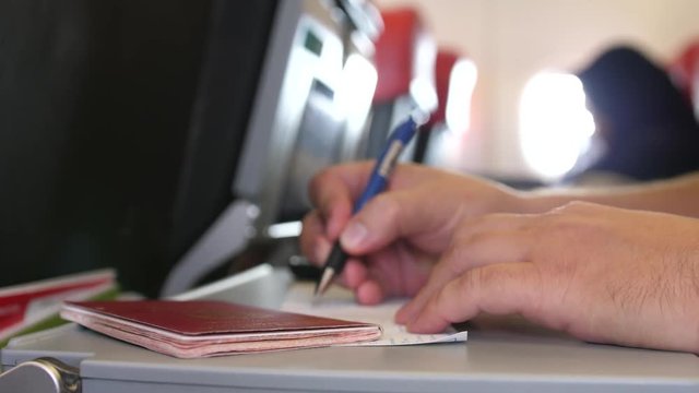 Close up of passenger with passport is filling in the migration or arrival cards in the plane while flight. 3840x2160