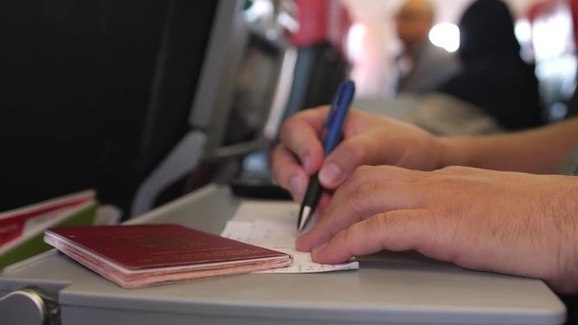 Close up of male hands passenger with passport is filling in the migration or arrival cards in the plane while flight. 3840x2160