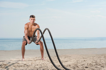 Cross power training. Young shirtless muscular man training with battle ropes on the beach against...