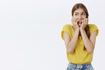 Fototapeta premium Waist-up shot of concerned nervous and anxious cute young timid woman in yellow t-shirt biting fingernails looking scared at upper left corner frightened, overreacting posing against white background