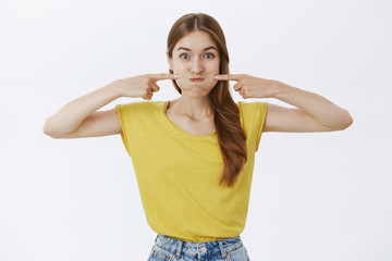 Fototapeta premium Charismatic funny and happy charming young female in yellow t-shirt pouting poking cheeks with index fingers and smiling as if holding breath playfully fooling around over white background