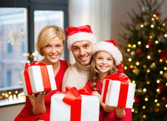 holidays, family and celebration concept - happy mother, father and daughter with gifts at home over christmas tree lights background