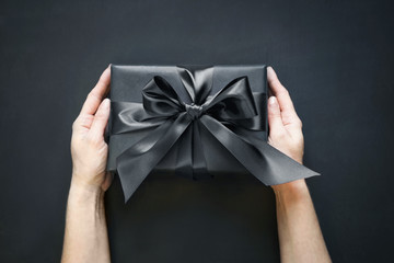 Gift box wrapped in black in female hand on black surface. Top view.