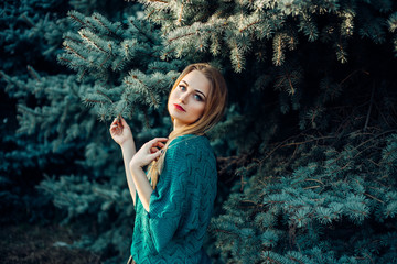 A girl with blond hair and blue eyes in a green sweater in a park near the green blue fir tree