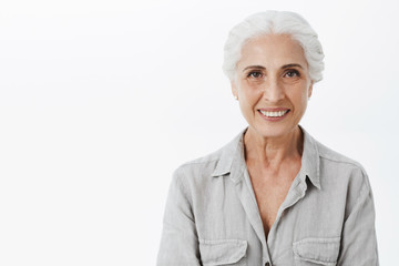 Waist-up shot of cute and kind wise elderly woman with white hair in casual shirt smiling broadly with assured and delighted look being amused and charismatic posing against gray background