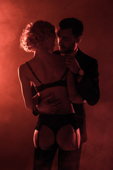 Back view of seductive woman in lingerie looking at man in costume on red smoke background