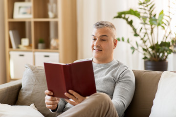 leisure, literature and people concept - man sitting on sofa and reading book at home